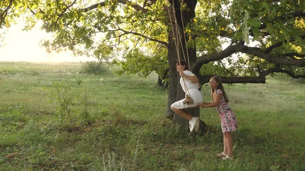Anak-anak berayun di dahan pohon oak di bawah sinar matahari. Mimpi untuk terbang. konsep masa kecil yang bahagia. Gadis-gadis cantik bermain di taman. gadis remaja menikmati terbang pada ayunan pada malam musim panas di hutan — Stok Foto