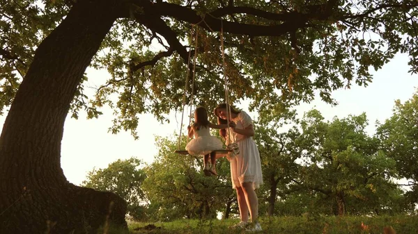 Mamma skakar en frisk liten dotter när hon svingar sig under träd i solen. mor leker med barn de svänger på ett rep på en ekgren i skogen. Flickan skrattar, jublar. Gratis familj som har kul i parken — Stockfoto