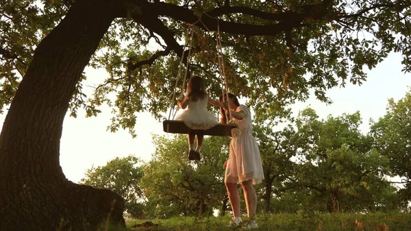Mamma skakar en frisk liten dotter när hon svingar sig under träd i solen. mor leker med barn de svänger på ett rep på en ekgren i skogen. Flickan skrattar, jublar. Gratis familj som har kul i parken — Stockfoto