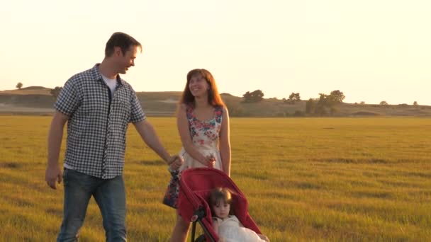 Caminar con un niño pequeño en cochecito en la naturaleza. familia feliz camina con su hija pequeña en un campo en el prado al atardecer. hija pequeña paseos en cochecito en el parque en el fondo del sol. concepto infantil — Vídeos de Stock