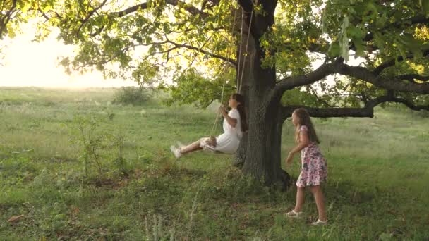 太陽の下でオークの木の枝でスイングしている子供たち空飛ぶ夢。幸せな子供時代の概念です美しい女の子たちが公園で遊んでいる。10代の女の子は森の中の夏の夜に飛び跳ねるのを楽しむ — ストック動画