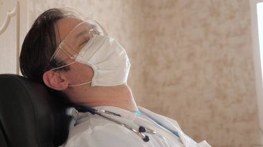 Klinikteki koruyucu maskeli ve gözlüklü yorgun doktor dinleniyor. Erkek doktor hastanede bir sandalyede uyuyakalmış. Sıhhiyeci gözlerini yorgunluktan ve fazla çalışmaktan kapattı.