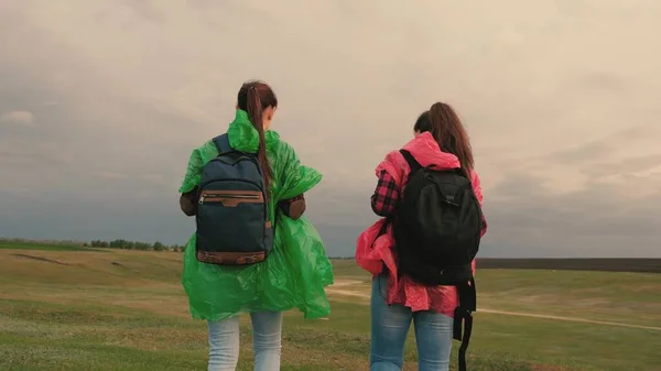Friska kvinnor turister reser med ryggsäckar i färgglada regnrockar. Sakta i backarna. Lagarbete för resenärer. Fria flickor-resenärer går till kanten av backen, glädjas, klappa händerna och hoppa med glädje. — Stockfoto