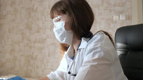 Телемедицина, концепція дистанційної медичної допомоги. Жінка-лікар у білому пальто та захисних окулярах, що працюють на ноутбуці, онлайн-консультації та конференції. Доктор спілкується з клієнтом за допомогою віртуального — стокове фото