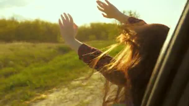 Volná žena cestuje autem chytí vítr s rukou z okna auta. Dívka s dlouhými vlasy sedí na předním sedadle auta, natahuje ruku ven oknem a oslnivě září zapadajícím sluncem — Stock video