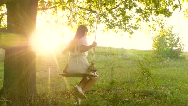 Jong meisje swingend op een schommel op een eiken tak in de zon. Dromen van vliegen. Gelukkige kindertijd concept. Mooi meisje in een witte jurk in het park. tiener meisje geniet van vliegen op swing op zomeravond in bos — Stockvideo