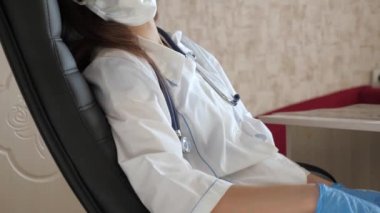 Sıhhiye yorgunluktan ve aşırı çalışmaktan gözlerini kapadı. Kadın doktor yorgun, koruyucu tıbbi maske takıyor ve klinikteki gözlükler dinleniyor. Kadın doktor hastanede bir sandalyede uyuyakalmış..