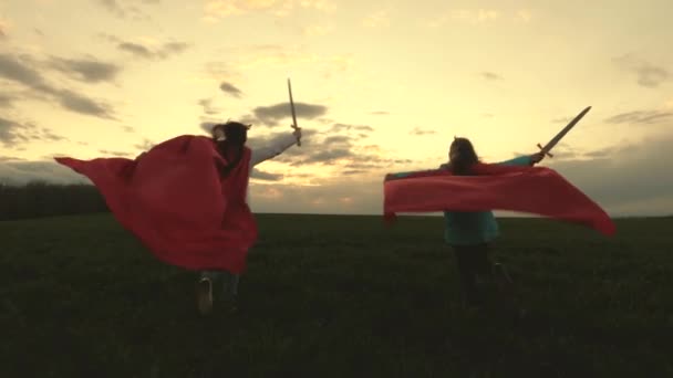 Meninas de manto vermelho correm com espadas na mão através do campo jogando cavaleiros medievais. As crianças lutam com uma espada de brinquedo. As crianças brincam aos cavaleiros. conceito de infância feliz. meninas saudáveis jogar super — Vídeo de Stock