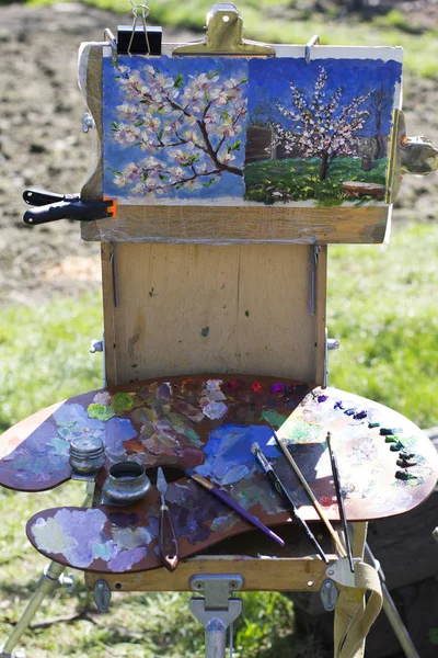 Plein air Etude with two paintings and a palette. A palette with multicolored paints and brushes lies on the sketch pad. Against the background of a spring green grass. Plenary, creativity, hobby, inspiration, harmony.