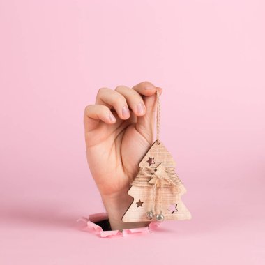 A male hand sticks out of a hole in pink paper, holds a Christmas tree ornament. Christmas concept symbol, commercial bucket print, copy space.