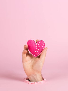 A male hand sticks out of a hole in pink paper, holds a handmade heart. Valentine's day concept, symbol of love, print commercial blank, copy space.