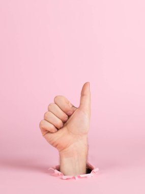 Male hand sticks out of a hole in paper, on a pink background. Pointing sign thumbs up, copy space.