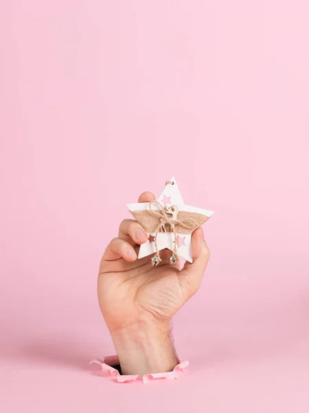 Eine männliche Hand ragt aus einem Loch in rosa Papier, hält einen Christbaumschmuck. weihnachtskonzept symbol, kommerzielle eimerdruck, kopierraum. — Stockfoto