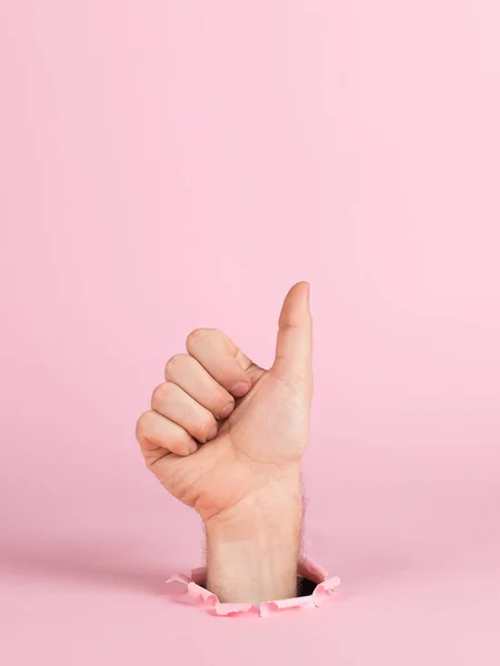 Männliche Hand ragt aus einem Loch in Papier, auf rosa Hintergrund. Zeigen Zeichen Daumen hoch, Kopierraum. — Stockfoto