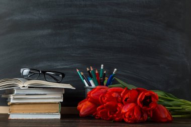 Kara tahta üzerinde kitaplar, kırmızı laleler ve okul malzemeleri. Öğretmenler günü için kartpostal konsepti. Boşluğu kopyala.