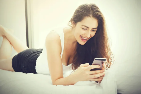 Beautiful Caucasian woman laying down on white bed using mobile phone and smile (Vintage tone)