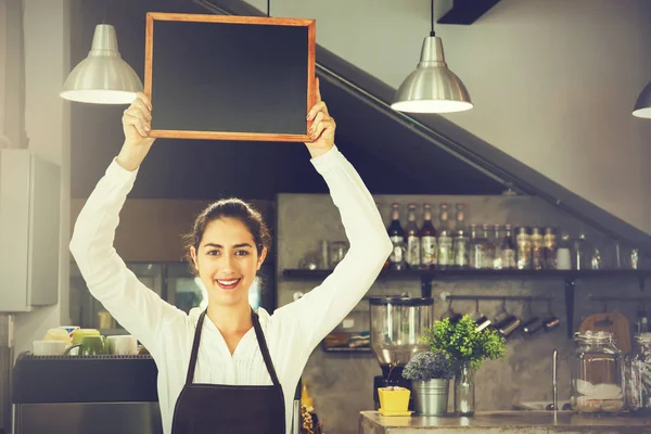 Vackra kaukasiska kvinna i barista förkläde med tomma blackboard skylt inuti coffee shop - redo att infoga text — Stockfoto