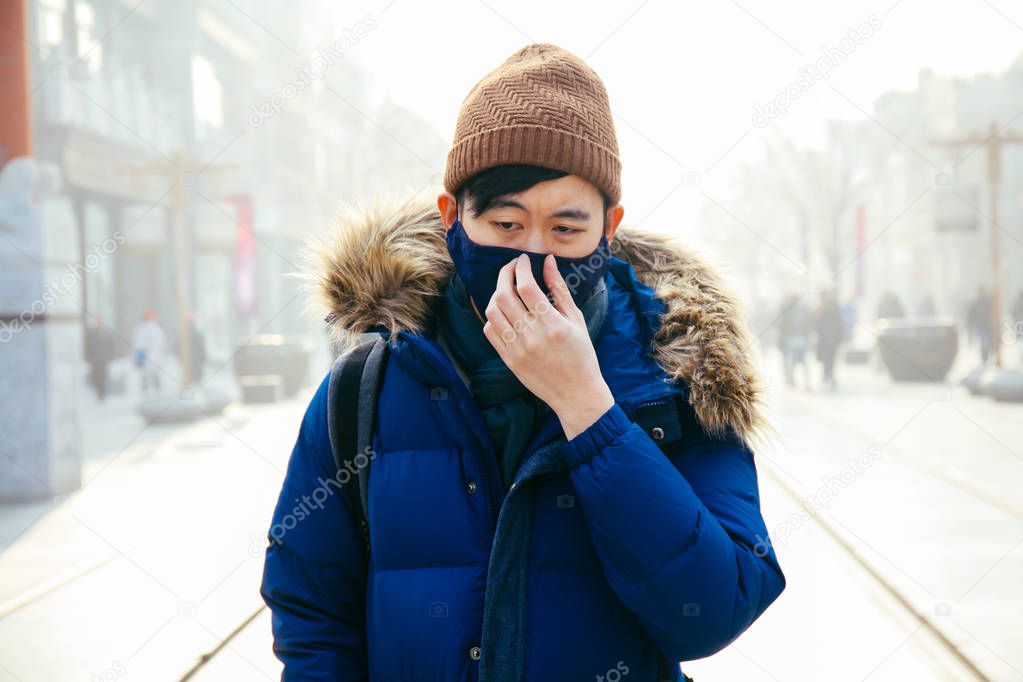 Asian man walking and wearing a face mask in a foggy smog and hazy day as he suffers from severe air pollution in Beijing, China 