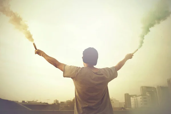 Manliga tonåringen holding färgglada röken sticker upp i luften över urbana staden bakgrund — Stockfoto