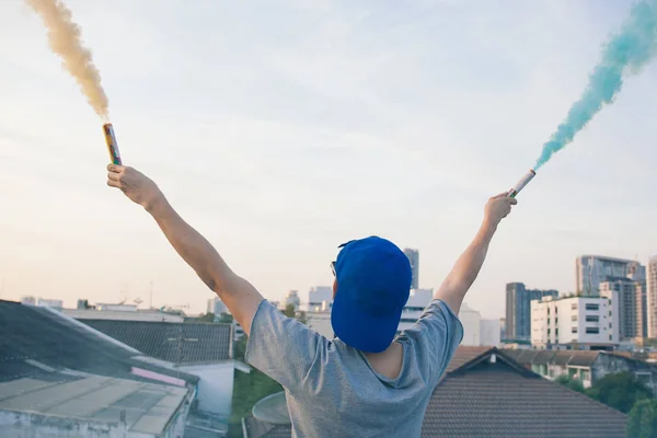 Manliga tonåringen holding färgglada röken sticker upp i luften över urbana staden bakgrund — Stockfoto