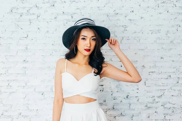 Молодая счастливая азиатка улыбается в модном платье и летнюю шляпу над белой кирпичной стеной — стоковое фото