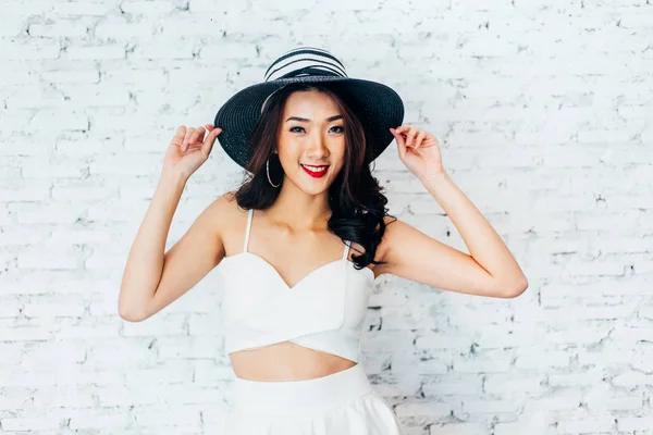 Молодая счастливая азиатка улыбается в модном платье и летнюю шляпу над белой кирпичной стеной — стоковое фото