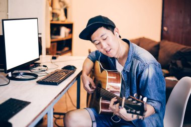 Genç Asyalı adam bilgisayar monitöründe gitar çalmayı öğrenme. Erkek gitaristi izlerken çevrimiçi öğretici. Kırpma yolunu monitörde ekleyin