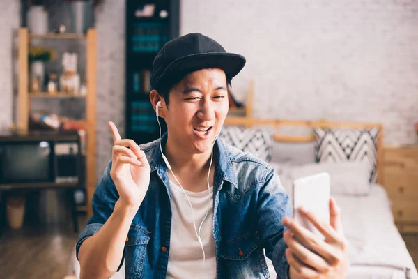 Young happy Asian man talking video call via smartphone wearing headphones at home — Stock Photo, Image