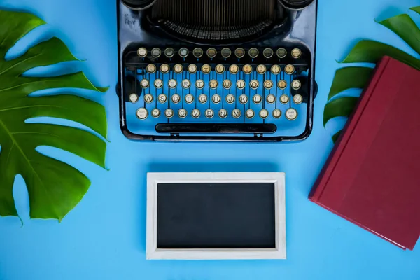 Old and vintage type writer machine and books and green leaves over blue background - with copy space on empty blackboard