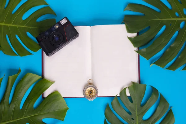 Ovanifrån - öppen bok med tomma sidor dekorerad med grön monstera blad, kamera, och kompass över pastell blå bakgrund - med kopia utrymme - semester och turism planering koncept — Stockfoto