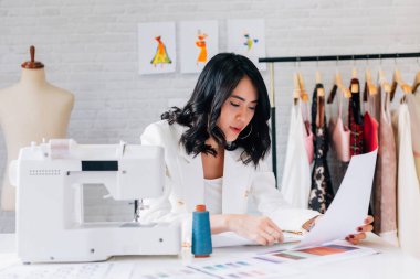 Genç Asyalı moda tasarımcısı dikiş makinesi ve giysi ekipmanıyla çizim ve renk çizelgesi üzerinde çalışıyor.