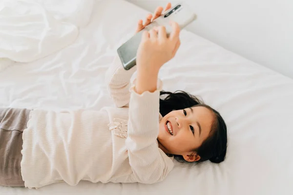 Joyful Asian child taking selfie on cellphone lying on bed — 스톡 사진