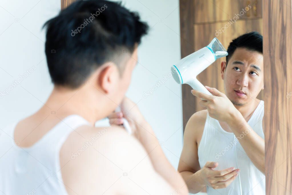 Back view of Asian guy in white shirt blowing dry hair using hairdryer in front of mirror