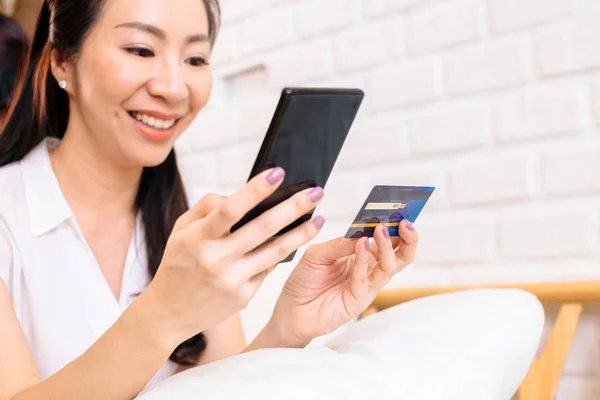 Rahat beyaz odada kredi kartı ve cep telefonu tutan 40s orta yaşlı Asyalı kadın Yüzü — Stok fotoğraf