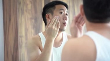 Genç Asyalı adam banyoda cilt kremi sürüyor.