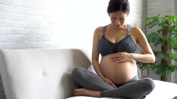 Jeune femme enceinte asiatique attend un nouvel enfant assis sur le canapé à la maison — Video