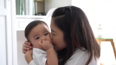Asian young adult mother kissing joyful son in living room.