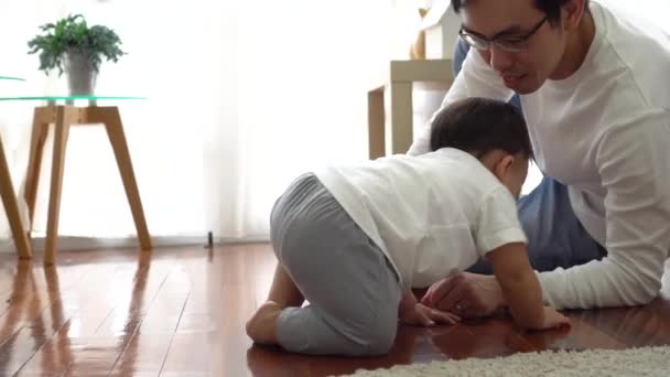Poważny Azji mały chłopiec czołga się na podłodze podczas gdy młody ojciec siedzi obok niego w salonie — Wideo stockowe