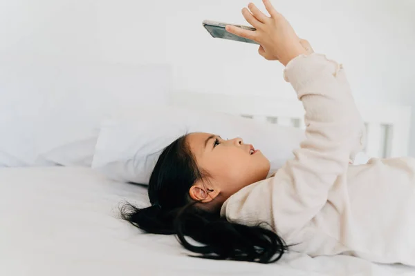 Спокойный азиатский ребенок играет в игры на сотовом телефоне лежит на кровати — стоковое фото