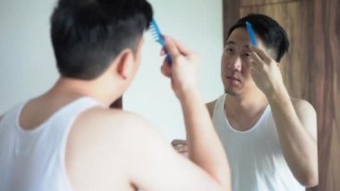 Genç bir erkek banyoda aynaya bakarken saçlarını tarıyor.