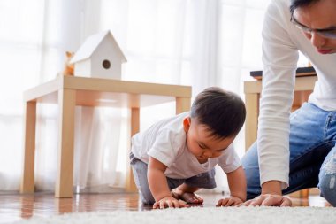 Serious Asian little boy crawling on floor while young father sitting next to him in living room