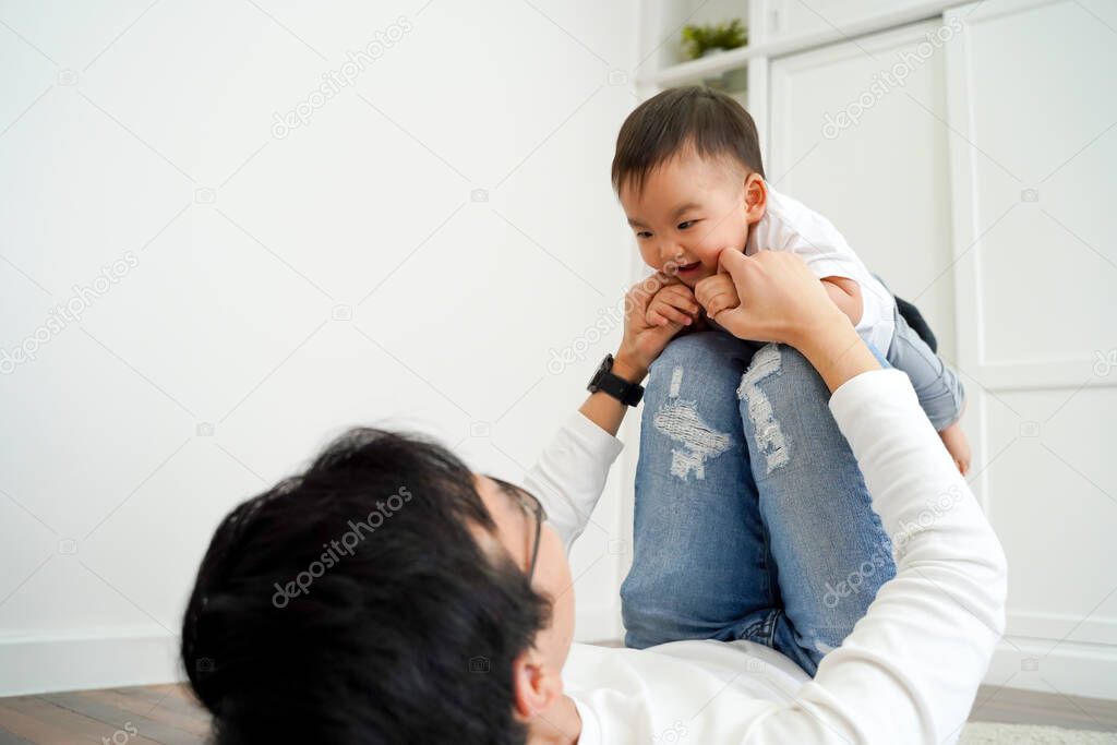 Joyful Asian boy playing airplane with parenting dad in the living room at home. Father is carrying innocent son up on his legs ready to fly. Fatherhood in Asia concept