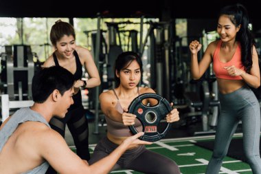 Bir grup insan, spor salonunda ağırlık plakasıyla çömelme hareketi yapan Asyalı bayan arkadaşına tezahürat yapıyor. Takım çalışması olarak çalışıyoruz..