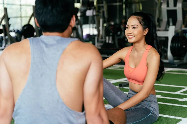 Gesicht einer jungen asiatischen, gemischt sportlichen Frau, die mit einem muskulösen Mann plaudert, der im Fitnessstudio auf dem Boden sitzt. — Stockfoto