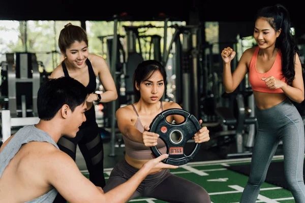 Gruppe von Menschen, die ihre asiatische Freundin bei Kniebeugen mit einer Hantelscheibe im Fitnessstudio anfeuern. Gemeinsam als Team arbeiten. — Stockfoto