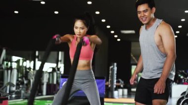 Asyalı kişisel antrenör vücut geliştiren bir kadına fitness salonunda çapraz dövüş halatları egzersizi yaptırıyor.