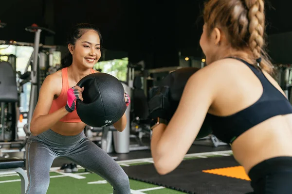 Glückliche junge Sportlerinnen, die mit Freunden Kniebeuge-Übungen machen und im Fitnessstudio einen Medizinball in der Hand halten. Gruppe von zwei selbstbewussten Frauen mit gesundem Lebensstil, die zusammen trainieren. — Stockfoto