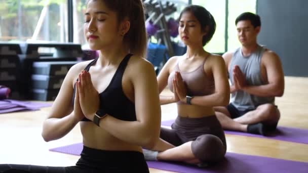 Grupo de mujeres asiáticas y hombres haciendo pilates tumbados en colchonetas de yoga en clase de aeróbic. Jóvenes deportistas en piernas cruzadas en el suelo y hacen namaste pose en gimnasio estudio — Vídeo de stock