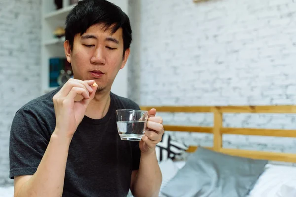 Молодой азиат чувствует себя больным и принимает таблетки, сидя дома на кровати — стоковое фото