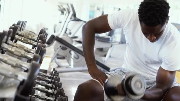Joven afroamericano sentado y levantando una mancuerna cerca del estante en el gimnasio. Hombre entrenamiento con pesas persona haciendo un curl bíceps en el gimnasio — Vídeos de Stock